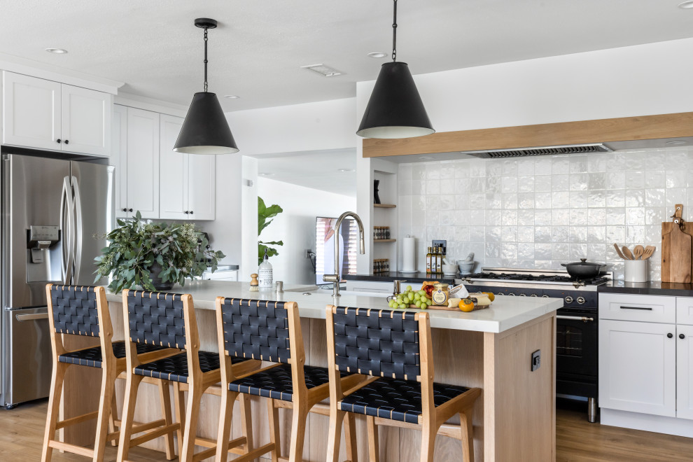 This is an example of a country l-shaped kitchen in Los Angeles with a farmhouse sink, shaker cabinets, white cabinets, white splashback, black appliances, medium hardwood floors, with island, brown floor and grey benchtop.
