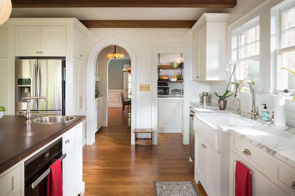 Example of a mid-sized transitional medium tone wood floor and brown floor kitchen pantry design in Atlanta with shaker cabinets, white cabinets, white backsplash, subway tile backsplash, stainless steel appliances, an island, a farmhouse sink, marble countertops and white countertops
