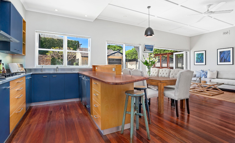 Photo of a mid-sized transitional u-shaped eat-in kitchen in Sydney with a double-bowl sink, blue cabinets, wood benchtops, blue splashback, glass sheet splashback, stainless steel appliances and medium hardwood floors.