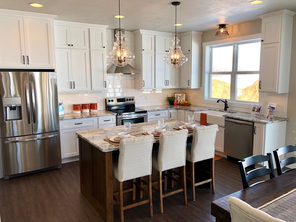 This is an example of a large country l-shaped eat-in kitchen in Other with a farmhouse sink, shaker cabinets, white cabinets, granite benchtops, white splashback, subway tile splashback, stainless steel appliances, vinyl floors, with island, brown floor and multi-coloured benchtop.