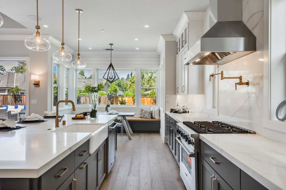 This is an example of a large country open plan kitchen in San Francisco with a farmhouse sink, shaker cabinets, grey cabinets, quartz benchtops, white splashback, engineered quartz splashback, stainless steel appliances, light hardwood floors, with island, grey floor and white benchtop.