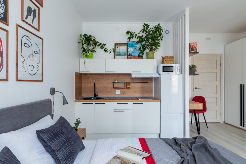 This is an example of a small contemporary single-wall open plan kitchen in Saint Petersburg with an integrated sink, flat-panel cabinets, white cabinets, wood benchtops, timber splashback, white appliances and light hardwood floors.