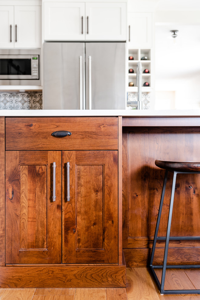 Example of a mid-sized transitional l-shaped medium tone wood floor and brown floor kitchen pantry design with an undermount sink, shaker cabinets, white cabinets, quartzite countertops, gray backsplash, porcelain backsplash, stainless steel appliances, an island and white countertops