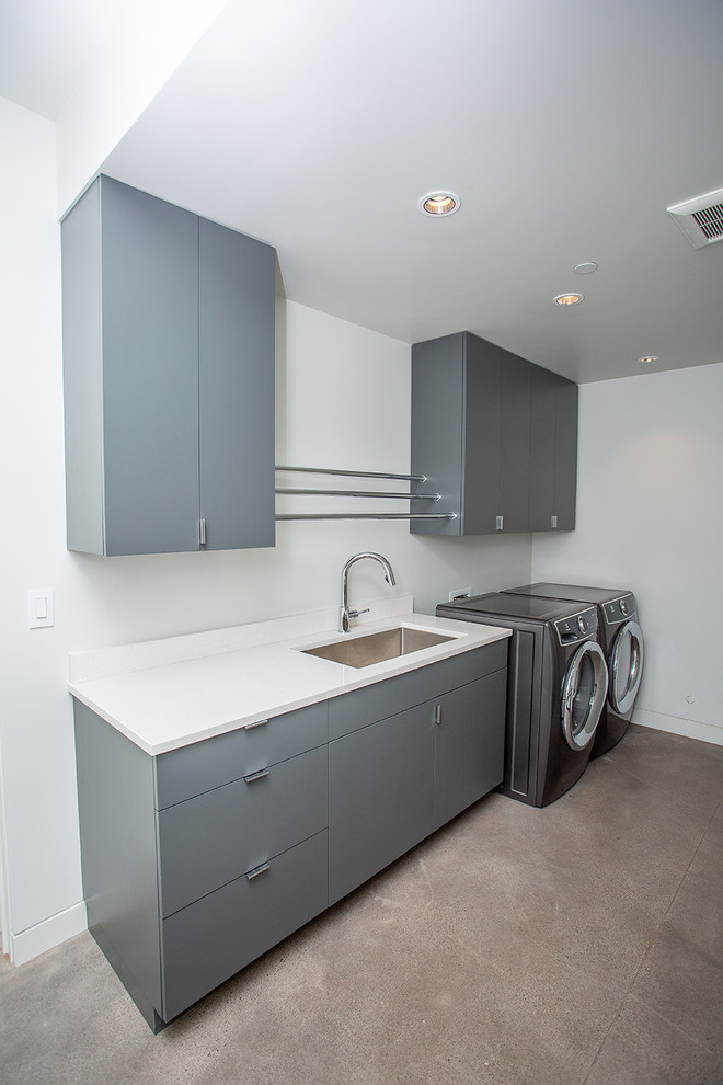 Photo of a mid-sized midcentury single-wall dedicated laundry room in Portland with an undermount sink, flat-panel cabinets, grey cabinets, white walls, concrete floors, a side-by-side washer and dryer, beige floor and white benchtop.