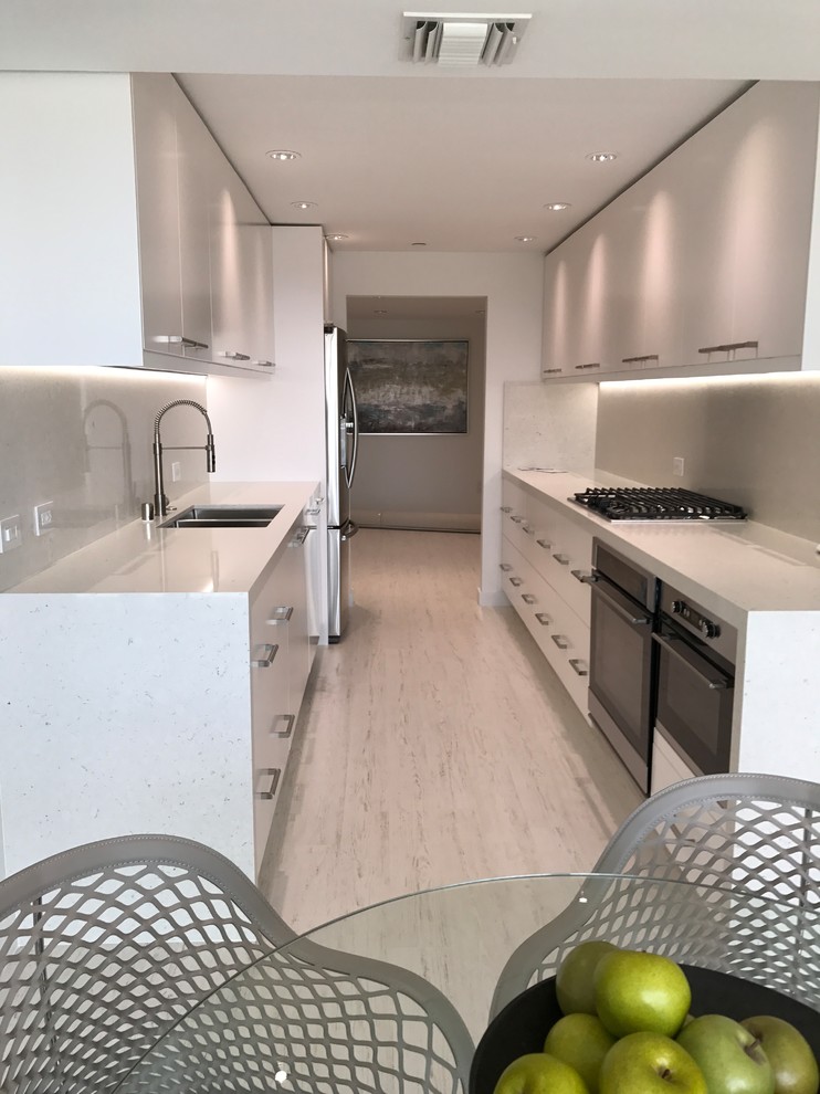 Photo of a mid-sized modern galley separate kitchen in Los Angeles with laminate floors, white floor, a double-bowl sink, flat-panel cabinets, white cabinets, quartz benchtops, white splashback, stone slab splashback, stainless steel appliances, no island and white benchtop.