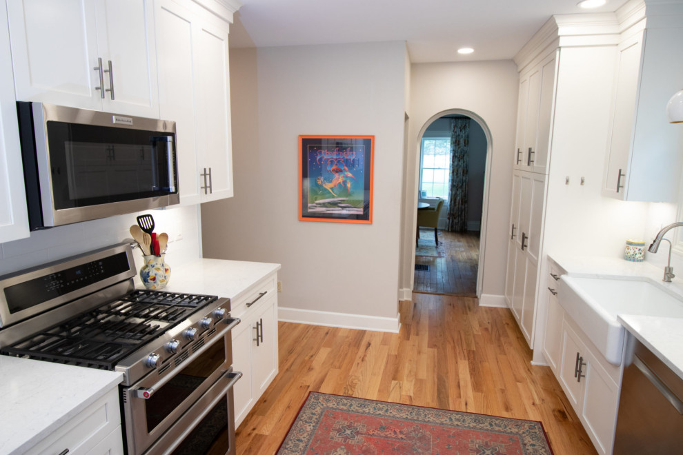 Example of a large transitional galley light wood floor and brown floor kitchen design with no island, white cabinets, quartz countertops, a farmhouse sink, white backsplash, glass tile backsplash, stainless steel appliances and white countertops