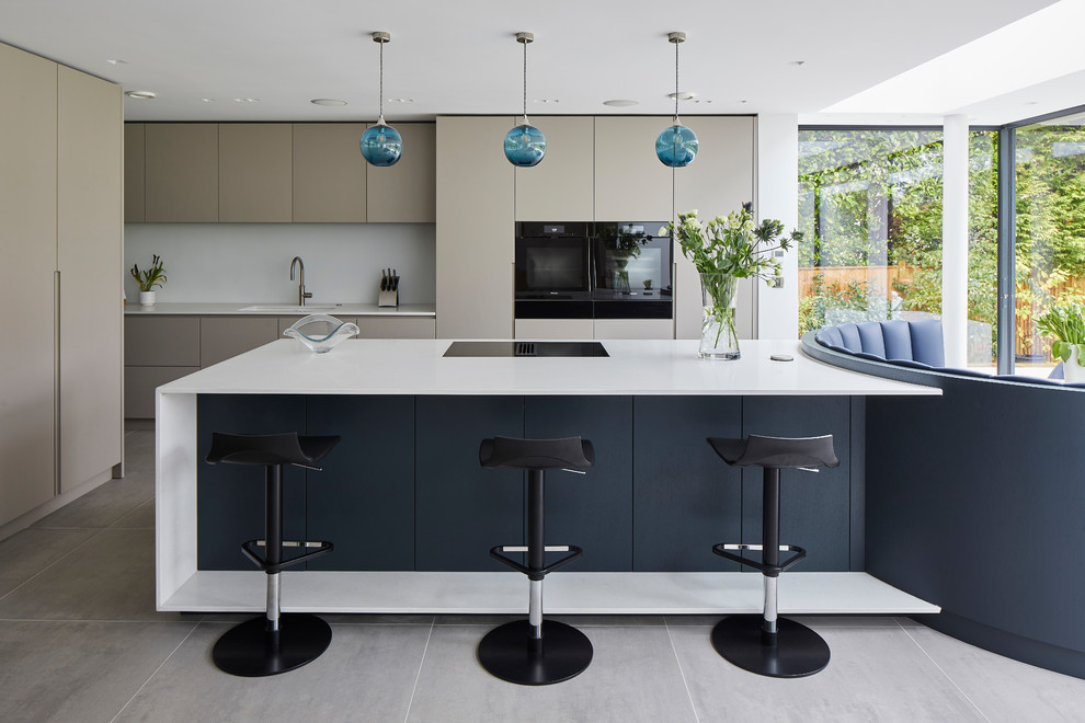 Contemporary galley open plan kitchen in Kent with an undermount sink, flat-panel cabinets, beige cabinets, white splashback, black appliances, with island, grey floor and white benchtop.