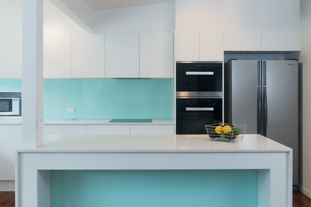 This is an example of a large beach style l-shaped eat-in kitchen in Brisbane with a drop-in sink, raised-panel cabinets, white cabinets, quartz benchtops, blue splashback, glass sheet splashback, stainless steel appliances, medium hardwood floors and with island.
