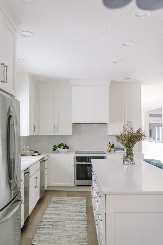 Photo of a large transitional l-shaped eat-in kitchen in Other with an undermount sink, shaker cabinets, white cabinets, quartz benchtops, white splashback, engineered quartz splashback, stainless steel appliances, laminate floors, with island, brown floor and white benchtop.