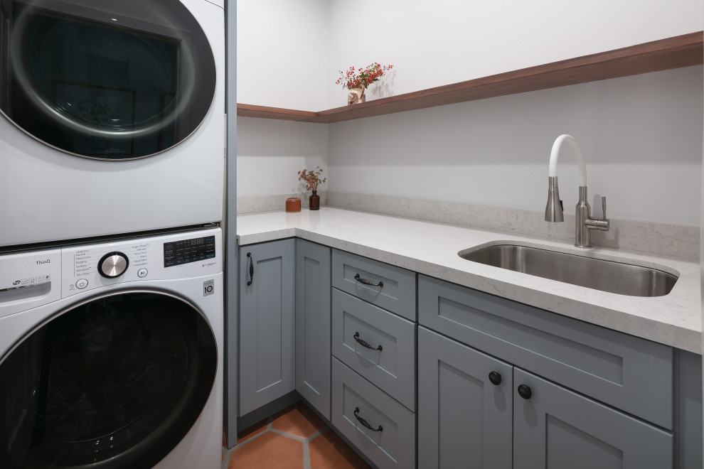 Inspiration for a small transitional l-shaped dedicated laundry room in Los Angeles with an undermount sink, shaker cabinets, grey cabinets, quartz benchtops, white splashback, stone slab splashback, white walls, terra-cotta floors, a stacked washer and dryer, orange floor and white benchtop.