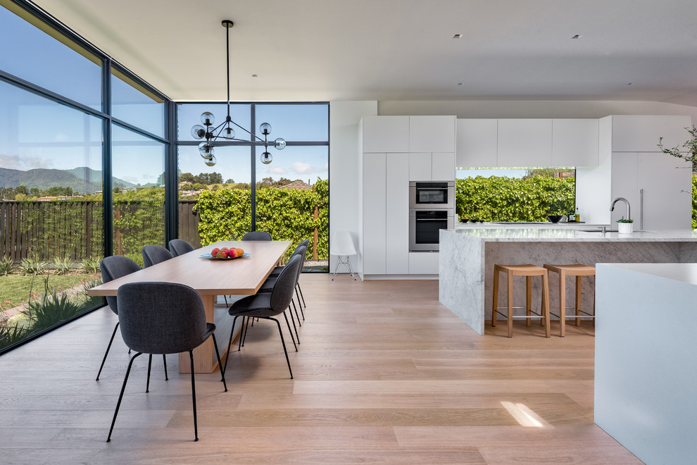 Large modern galley open plan kitchen in San Francisco with an undermount sink, flat-panel cabinets, white cabinets, marble benchtops, white splashback, marble splashback, stainless steel appliances, light hardwood floors, with island and white benchtop.