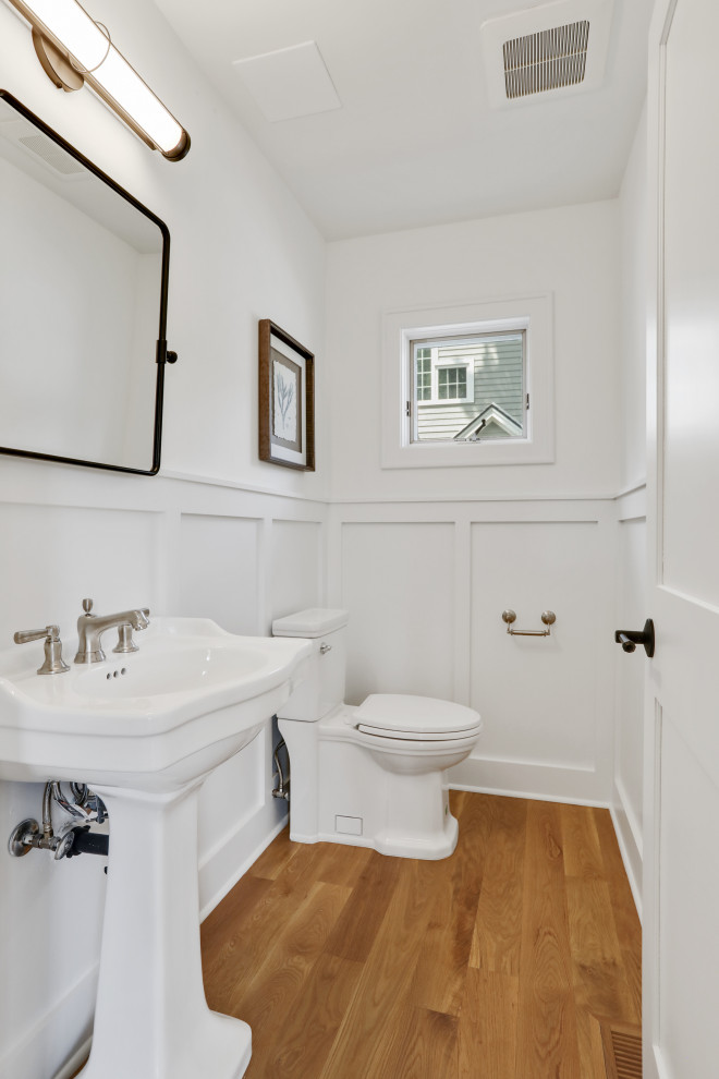 This is an example of a medium sized traditional cloakroom in Minneapolis with white cabinets, a two-piece toilet, white walls, light hardwood flooring, a pedestal sink, a freestanding vanity unit and panelled walls.