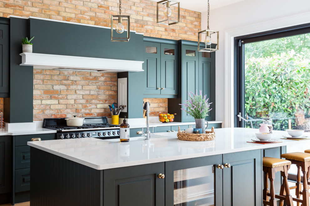 This is an example of a mid-sized transitional galley kitchen in Cork with a farmhouse sink, raised-panel cabinets, green cabinets, brick splashback, with island and white benchtop.