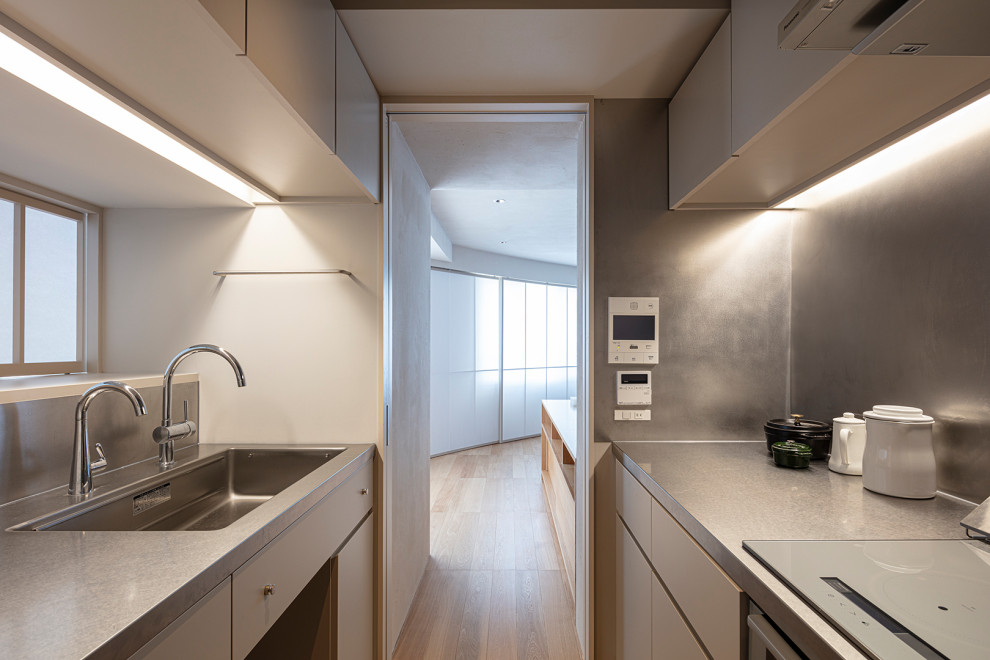 This is an example of a small modern galley separate kitchen in Tokyo with an integrated sink, white cabinets, stainless steel benchtops, stainless steel appliances, medium hardwood floors, no island and white benchtop.