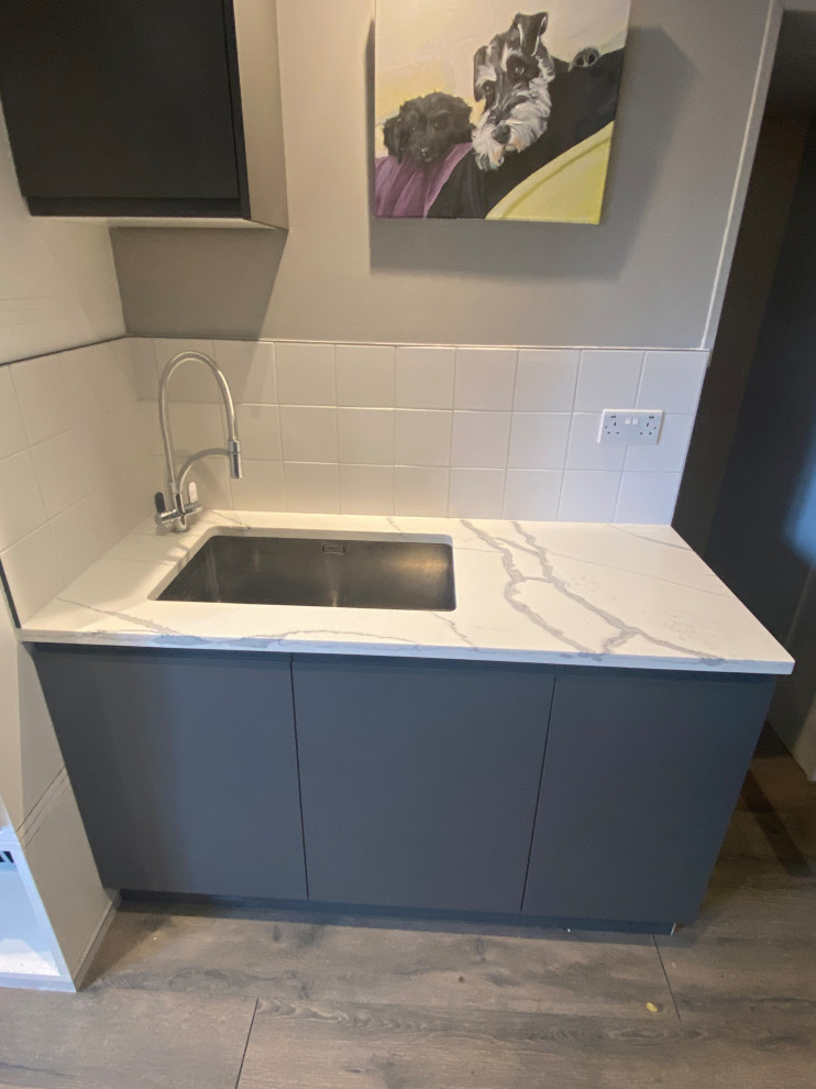Photo of a large contemporary galley utility room in London with a drop-in sink, flat-panel cabinets, grey cabinets, quartzite benchtops, white splashback, porcelain splashback, beige walls, laminate floors, a side-by-side washer and dryer, grey floor and multi-coloured benchtop.