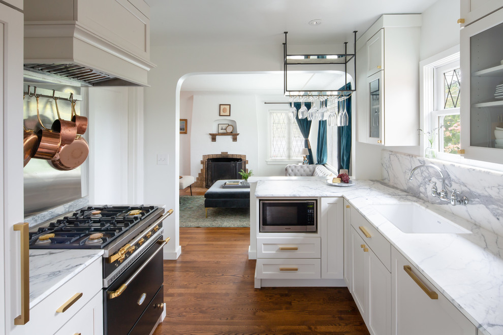This is an example of a small traditional galley open plan kitchen in Seattle with an undermount sink, shaker cabinets, beige cabinets, marble benchtops, white splashback, marble splashback, black appliances, medium hardwood floors, a peninsula, brown floor and white benchtop.