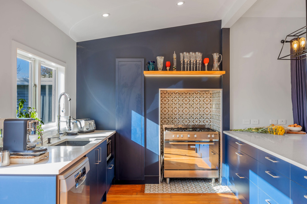 This is an example of a mid-sized eclectic u-shaped kitchen in Auckland with a double-bowl sink, flat-panel cabinets, blue cabinets, quartz benchtops, grey splashback, ceramic splashback, stainless steel appliances, medium hardwood floors, brown floor, white benchtop and coffered.