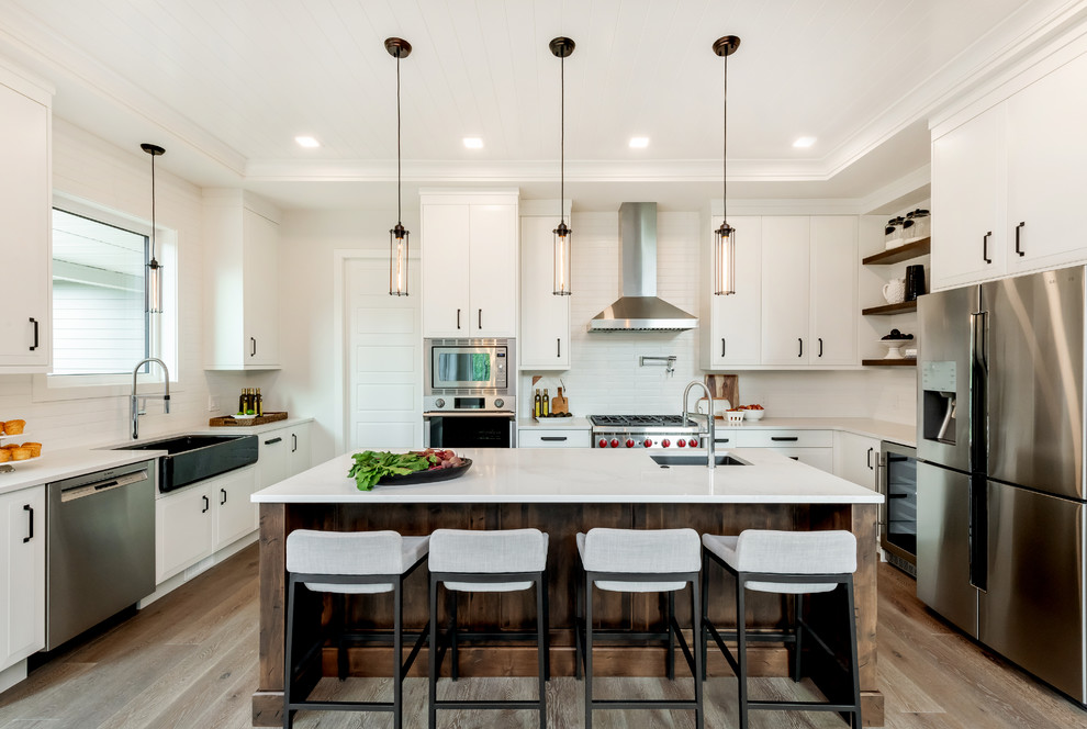 Photo of a large transitional u-shaped kitchen in Other with a farmhouse sink, white cabinets, quartz benchtops, white splashback, subway tile splashback, stainless steel appliances, medium hardwood floors, with island, brown floor and white benchtop.