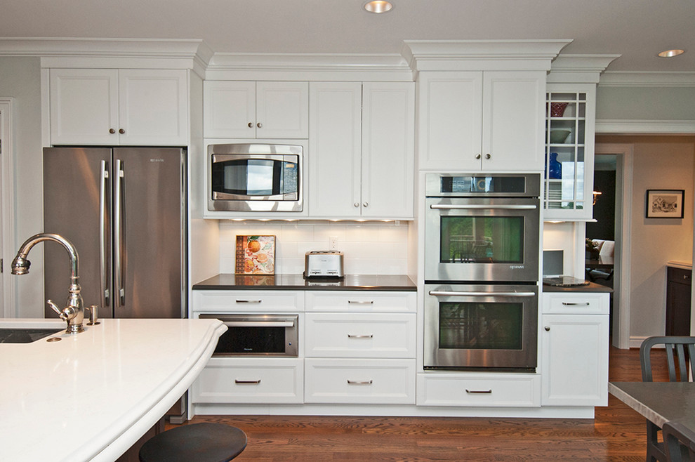 Example of a mid-sized transitional l-shaped medium tone wood floor and brown floor open concept kitchen design with an undermount sink, glass-front cabinets, white cabinets, white backsplash, subway tile backsplash, stainless steel appliances, an island and solid surface countertops