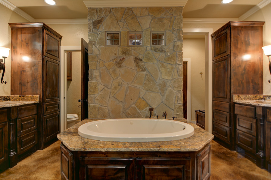 Drop-in bathtub - mid-sized rustic master brown tile and ceramic tile concrete floor drop-in bathtub idea with raised-panel cabinets, dark wood cabinets, a two-piece toilet, beige walls, granite countertops, an undermount sink, beige backsplash, stone tile backsplash, stainless steel appliances and an island