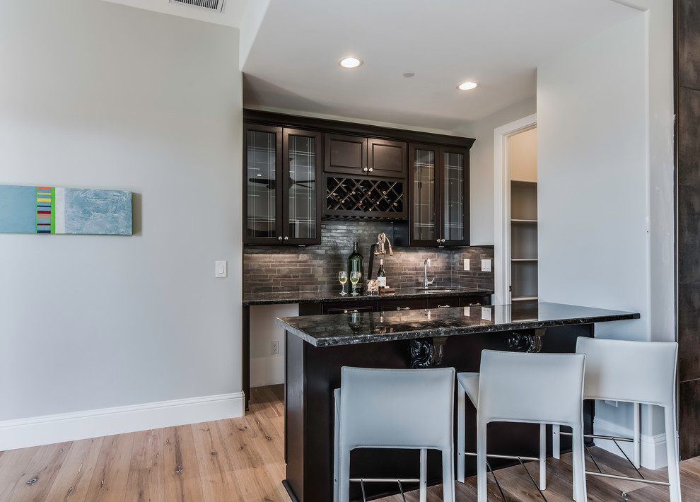 Small transitional galley seated home bar in Sacramento with an undermount sink, glass-front cabinets, dark wood cabinets, granite benchtops, black splashback, slate splashback, light hardwood floors and beige floor.