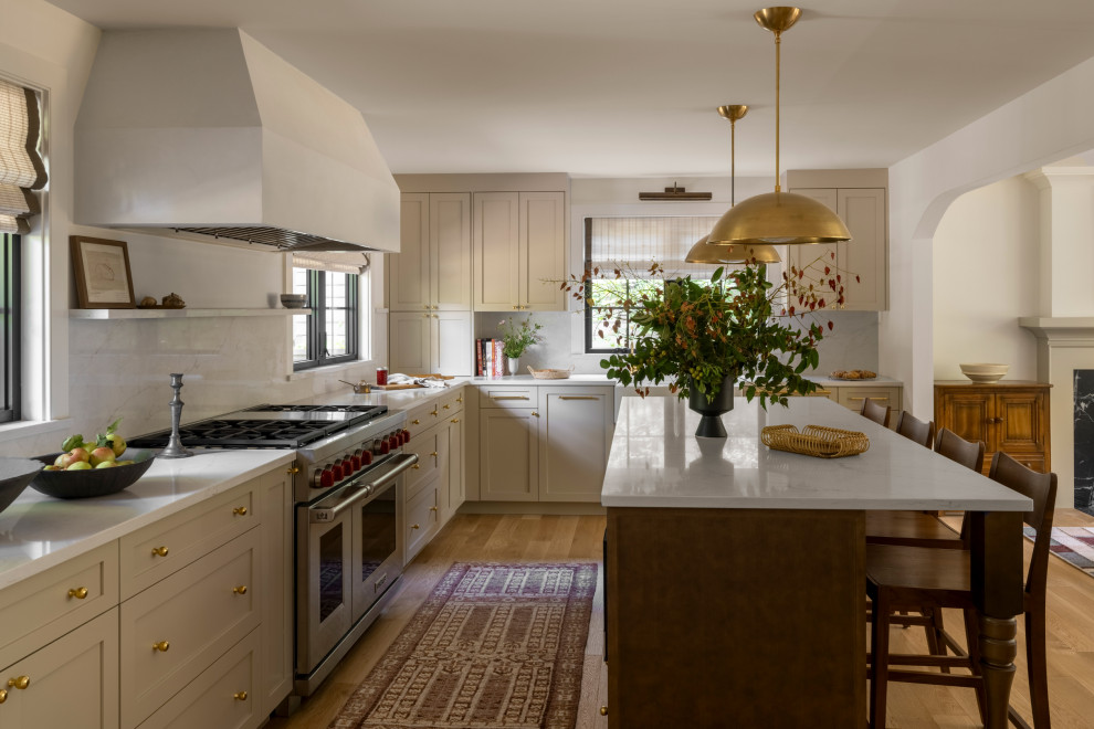 Example of a large classic l-shaped light wood floor and beige floor enclosed kitchen design in Seattle with a farmhouse sink, shaker cabinets, beige cabinets, marble countertops, white backsplash, stone slab backsplash, stainless steel appliances, white countertops and an island