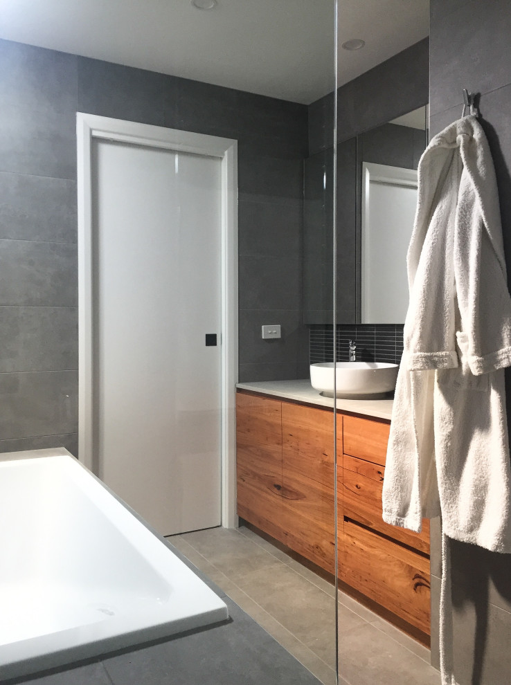 Inspiration for a medium sized contemporary ensuite bathroom in Melbourne with flat-panel cabinets, light wood cabinets, a built-in bath, a walk-in shower, grey tiles, ceramic tiles, grey walls, ceramic flooring, a vessel sink, engineered stone worktops, grey floors, an open shower, grey worktops, an enclosed toilet, a single sink and a built in vanity unit.