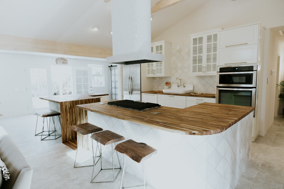 Inspiration for a mid-sized scandinavian u-shaped open plan kitchen in Phoenix with a farmhouse sink, glass-front cabinets, white cabinets, wood benchtops, white splashback, ceramic splashback, stainless steel appliances, porcelain floors, multiple islands, grey floor and brown benchtop.