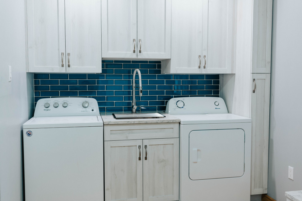 This is an example of a mid-sized transitional utility room in Toronto with an undermount sink, shaker cabinets, grey cabinets, quartz benchtops, blue splashback, glass tile splashback, grey walls, medium hardwood floors, a side-by-side washer and dryer, brown floor and grey benchtop.