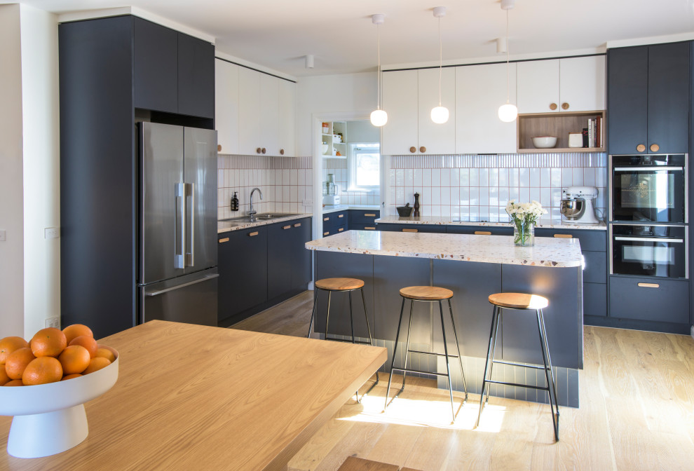 This is an example of a mid-sized midcentury l-shaped open plan kitchen in Melbourne with a double-bowl sink, terrazzo benchtops, subway tile splashback, black appliances, light hardwood floors, with island, flat-panel cabinets, blue cabinets, white splashback, beige floor and multi-coloured benchtop.