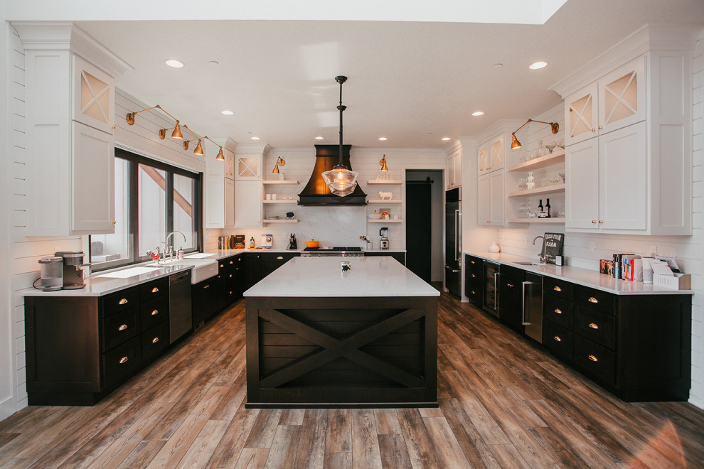 This is an example of a large country u-shaped eat-in kitchen in Portland with a farmhouse sink, recessed-panel cabinets, black cabinets, quartz benchtops, white splashback, subway tile splashback, stainless steel appliances, dark hardwood floors, with island, brown floor and white benchtop.