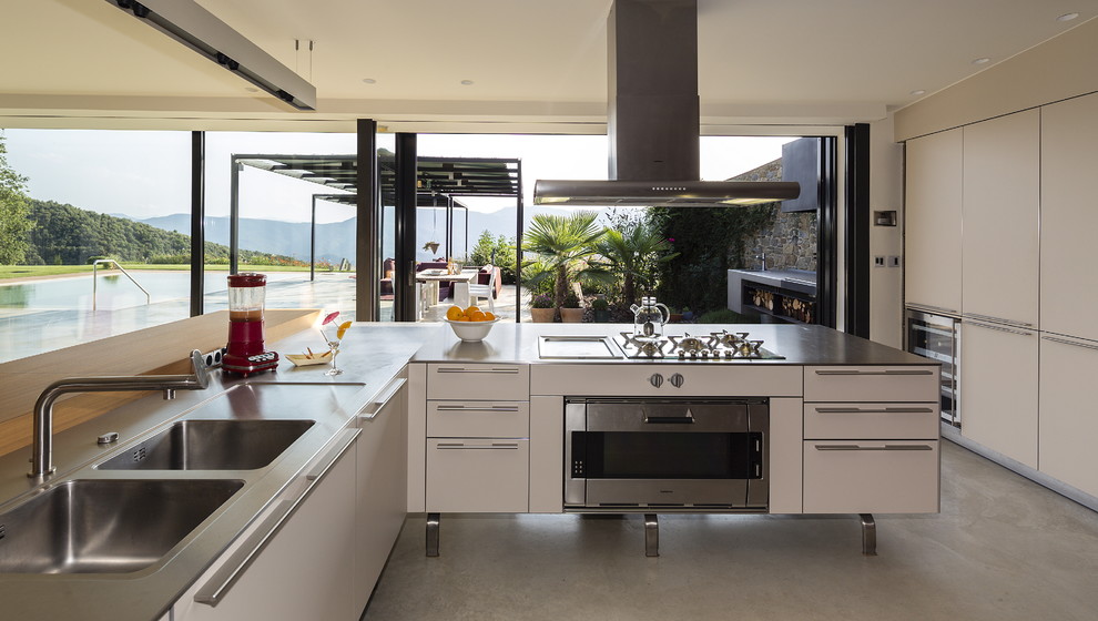 Photo of a large industrial l-shaped open plan kitchen in Barcelona with an integrated sink, flat-panel cabinets, white cabinets, stainless steel benchtops, stainless steel appliances, concrete floors and no island.