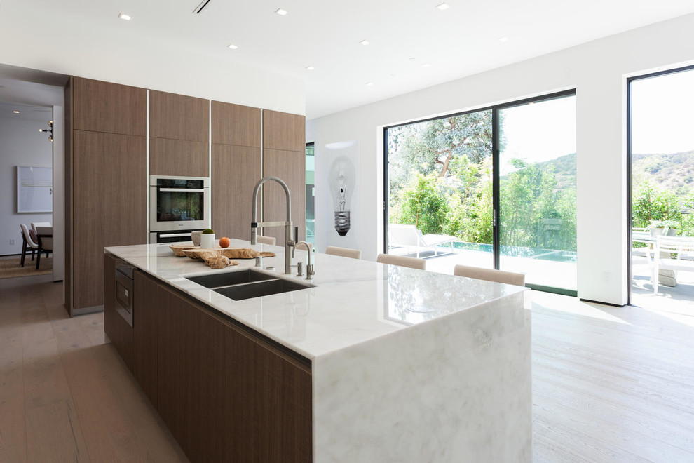 Photo of a large modern single-wall open plan kitchen in Los Angeles with a single-bowl sink, flat-panel cabinets, light wood cabinets, marble benchtops, white splashback, stone slab splashback, stainless steel appliances, light hardwood floors and with island.