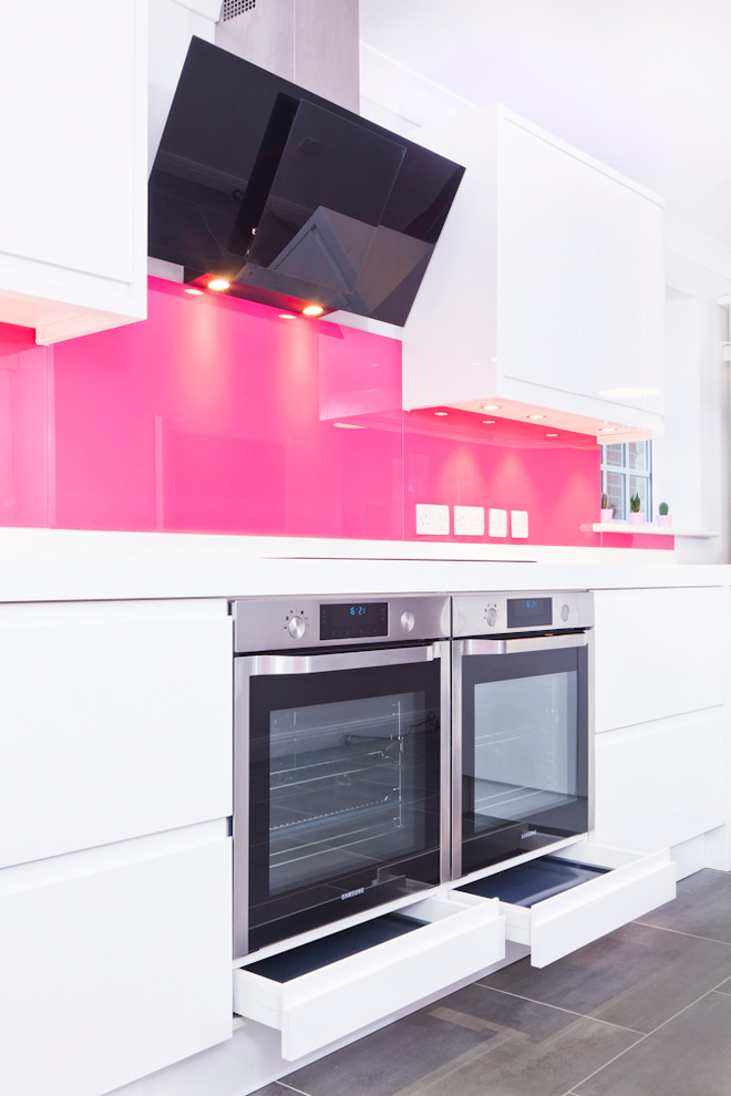 Mid-sized contemporary galley eat-in kitchen in Buckinghamshire with flat-panel cabinets, white cabinets, solid surface benchtops, pink splashback, glass sheet splashback, stainless steel appliances, vinyl floors, a peninsula and an integrated sink.