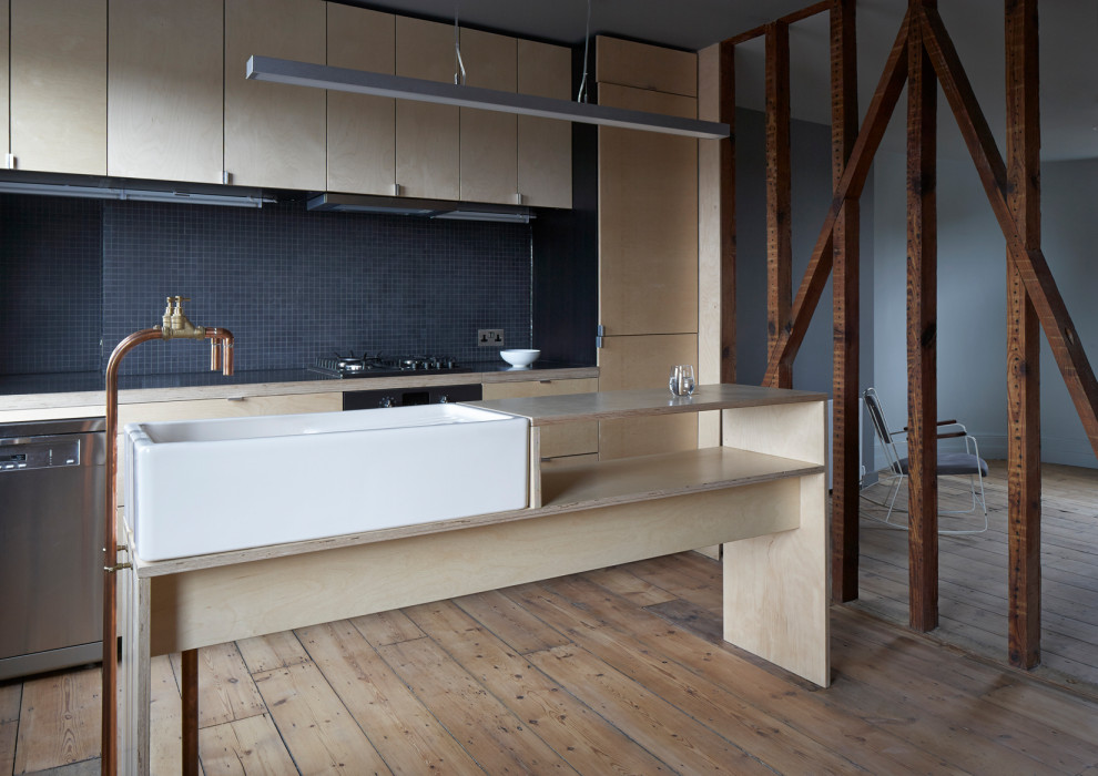 This is an example of an industrial open plan kitchen in London with a farmhouse sink, flat-panel cabinets, wood benchtops, black splashback, plywood floors, with island, light wood cabinets, mosaic tile splashback and stainless steel appliances.