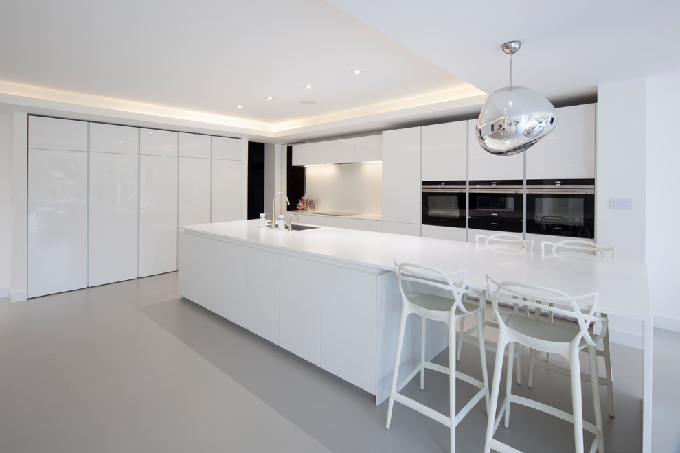 This is an example of an expansive contemporary l-shaped open plan kitchen in London with an integrated sink, flat-panel cabinets, white cabinets, concrete benchtops, white splashback, stainless steel appliances and a peninsula.