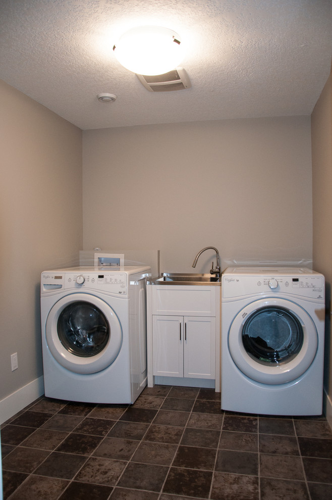This is an example of a mid-sized traditional single-wall dedicated laundry room in Calgary with an utility sink, shaker cabinets, white cabinets, stainless steel benchtops, grey walls, linoleum floors and a side-by-side washer and dryer.