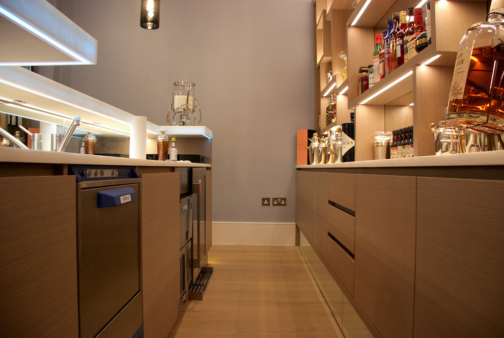 This is an example of a large modern single-wall bar cart in London with a drop-in sink, open cabinets, light wood cabinets, onyx benchtops, multi-coloured splashback, mirror splashback, light hardwood floors, grey floor and white benchtop.