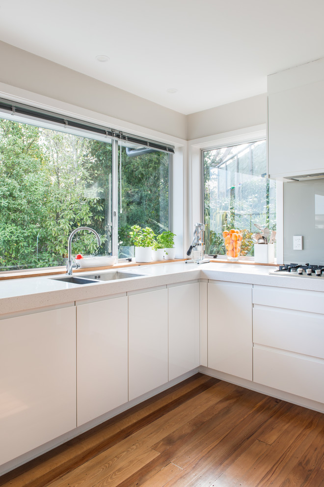 Mid-sized scandinavian u-shaped kitchen pantry in Christchurch with an undermount sink, flat-panel cabinets, white cabinets, solid surface benchtops, glass sheet splashback, white appliances, medium hardwood floors, a peninsula and brown floor.