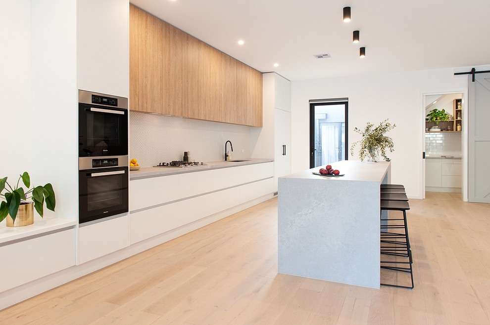Design ideas for an expansive modern galley open plan kitchen in Melbourne with an undermount sink, white cabinets, quartz benchtops, grey splashback, black appliances, light hardwood floors, with island, grey benchtop, flat-panel cabinets and beige floor.