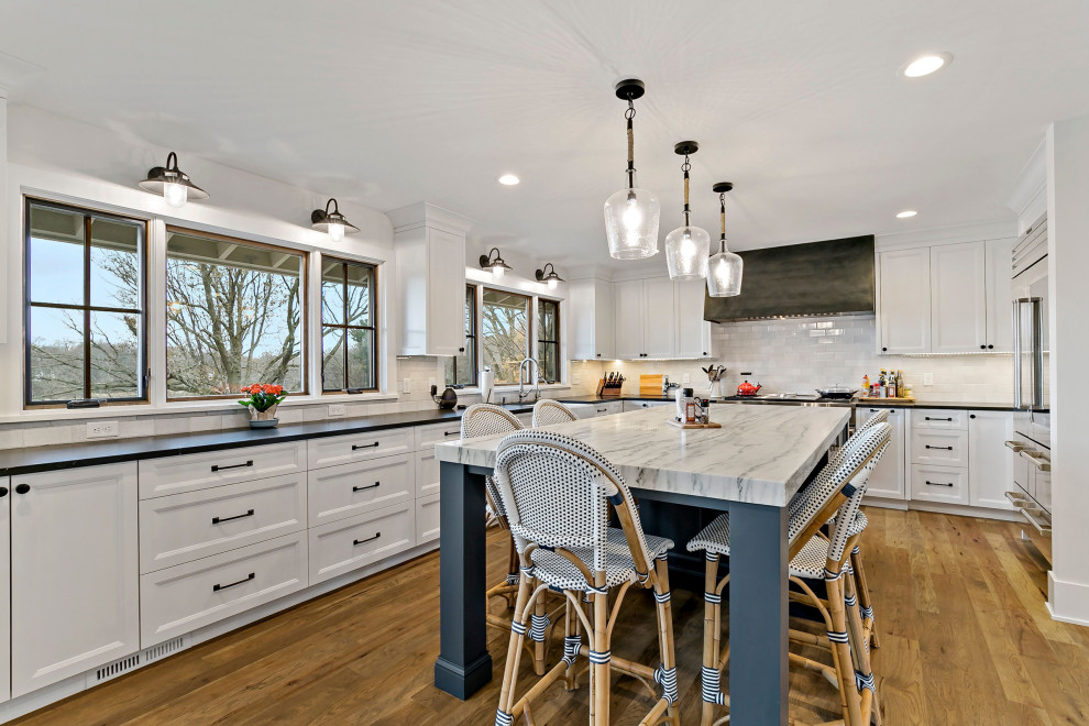 Photo of a large traditional u-shaped kitchen/diner in Minneapolis with a belfast sink, shaker cabinets, white cabinets, engineered stone countertops, white splashback, stainless steel appliances, medium hardwood flooring, an island and black worktops.