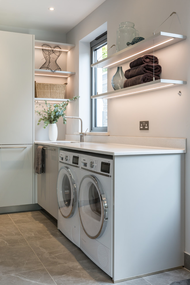 This is an example of a mid-sized modern u-shaped dedicated laundry room in Gloucestershire with a drop-in sink, flat-panel cabinets, grey cabinets, solid surface benchtops, white walls, ceramic floors, a side-by-side washer and dryer, brown floor and white benchtop.