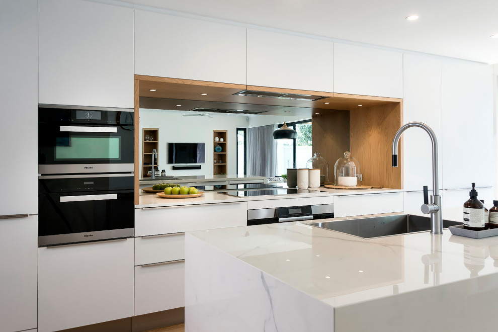 Design ideas for a large scandinavian galley open plan kitchen in Perth with a single-bowl sink, flat-panel cabinets, white cabinets, quartz benchtops, metallic splashback, mirror splashback, black appliances, light hardwood floors, with island, brown floor and white benchtop.