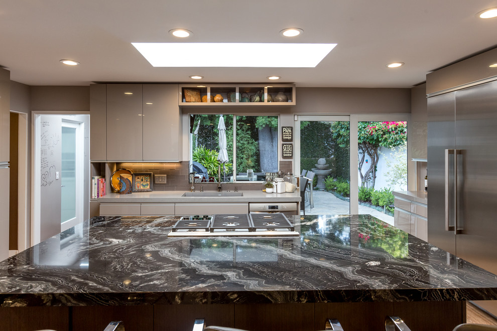 Design ideas for a large midcentury u-shaped open plan kitchen in Los Angeles with an undermount sink, flat-panel cabinets, grey cabinets, granite benchtops, grey splashback, glass tile splashback, stainless steel appliances, medium hardwood floors and with island.