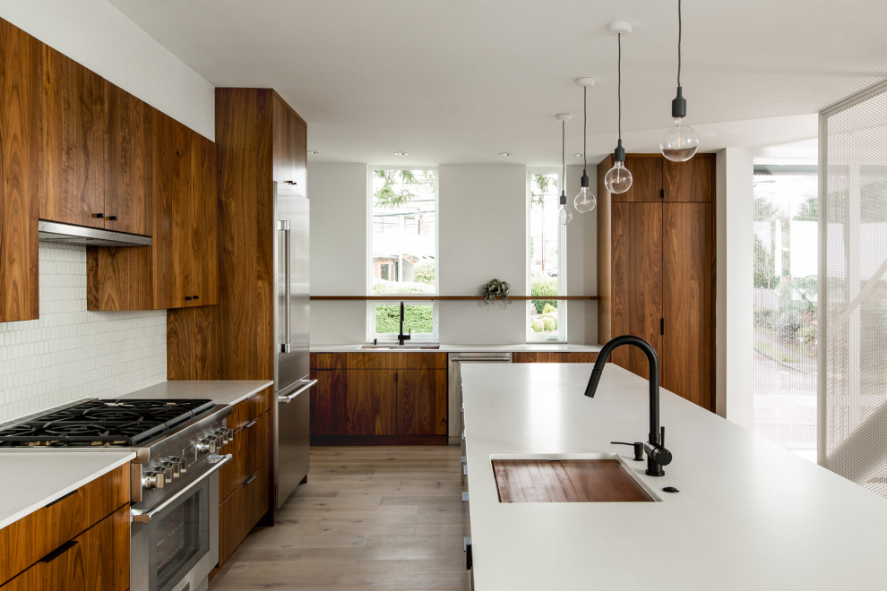 Large midcentury l-shaped kitchen in Seattle with a submerged sink, flat-panel cabinets, brown cabinets, engineered stone countertops, white splashback, ceramic splashback, stainless steel appliances, light hardwood flooring, an island and white worktops.