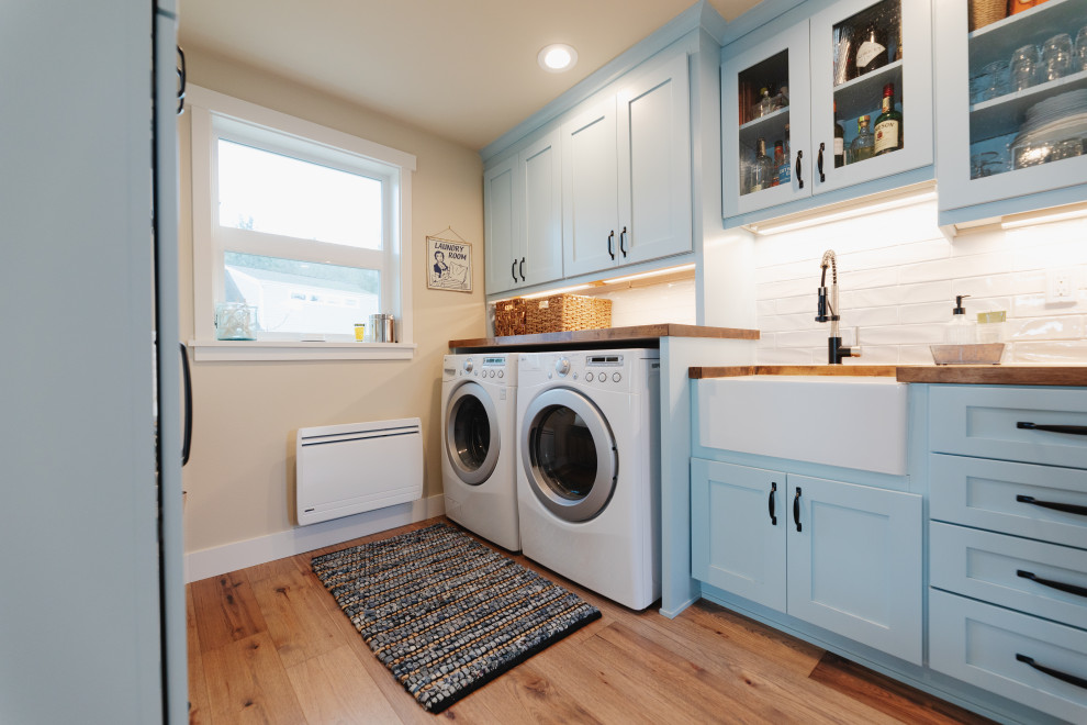 Large beach style galley utility room in Seattle with a farmhouse sink, shaker cabinets, blue cabinets, wood benchtops, white splashback, ceramic splashback, beige walls, medium hardwood floors, a side-by-side washer and dryer, brown floor and brown benchtop.