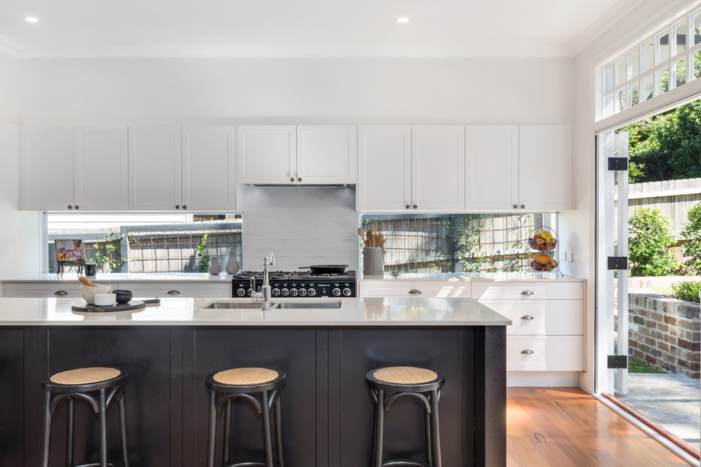 Transitional galley open plan kitchen in Sydney with a drop-in sink, shaker cabinets, quartz benchtops, glass sheet splashback, black appliances, light hardwood floors, with island and brown floor.