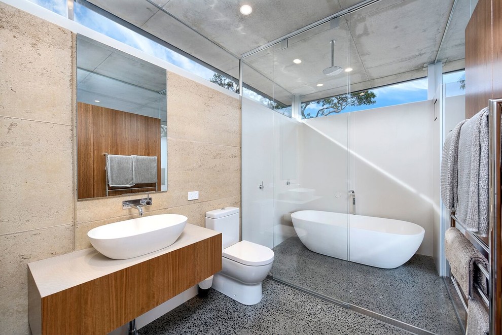 This is an example of a mid-sized contemporary master wet room bathroom in Sydney with flat-panel cabinets, brown cabinets, a drop-in tub, a two-piece toilet, ceramic tile, beige walls, limestone floors, a vessel sink, wood benchtops, grey floor, a sliding shower screen and brown benchtops.