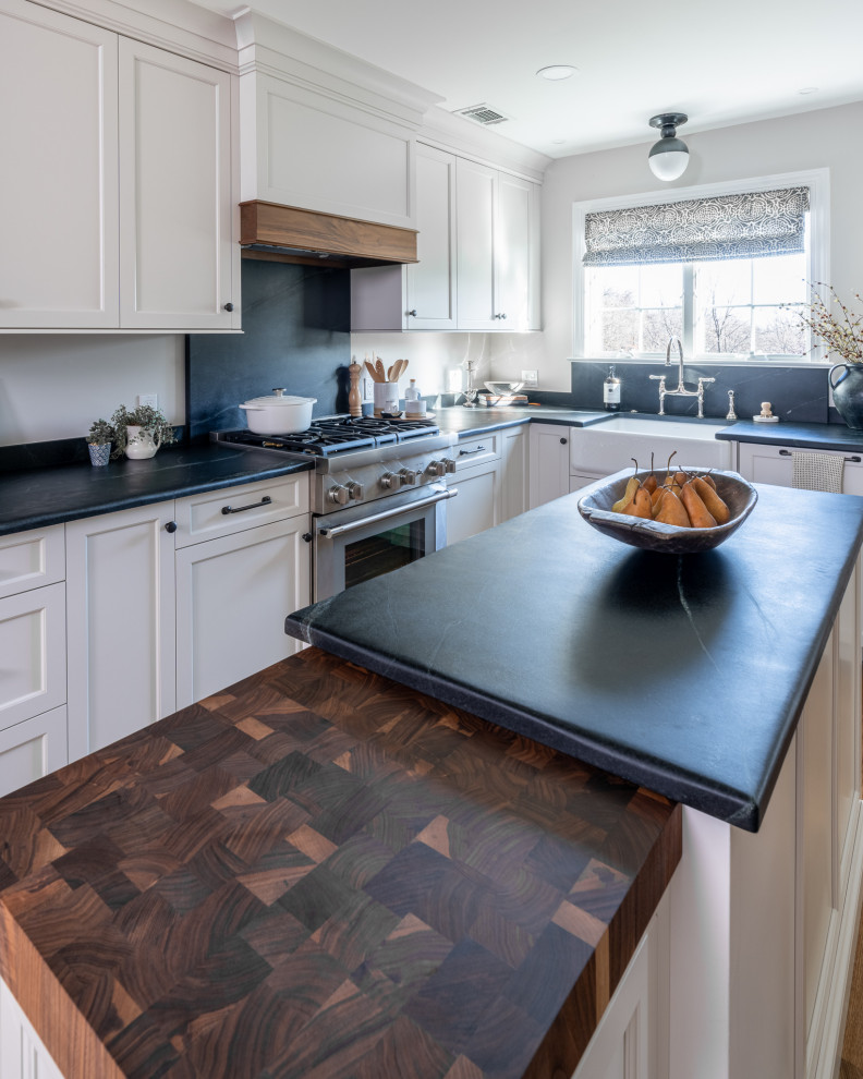 Mid-sized transitional l-shaped eat-in kitchen in Philadelphia with a farmhouse sink, shaker cabinets, grey cabinets, soapstone benchtops, black splashback, stone slab splashback, stainless steel appliances, medium hardwood floors, with island, brown floor and black benchtop.