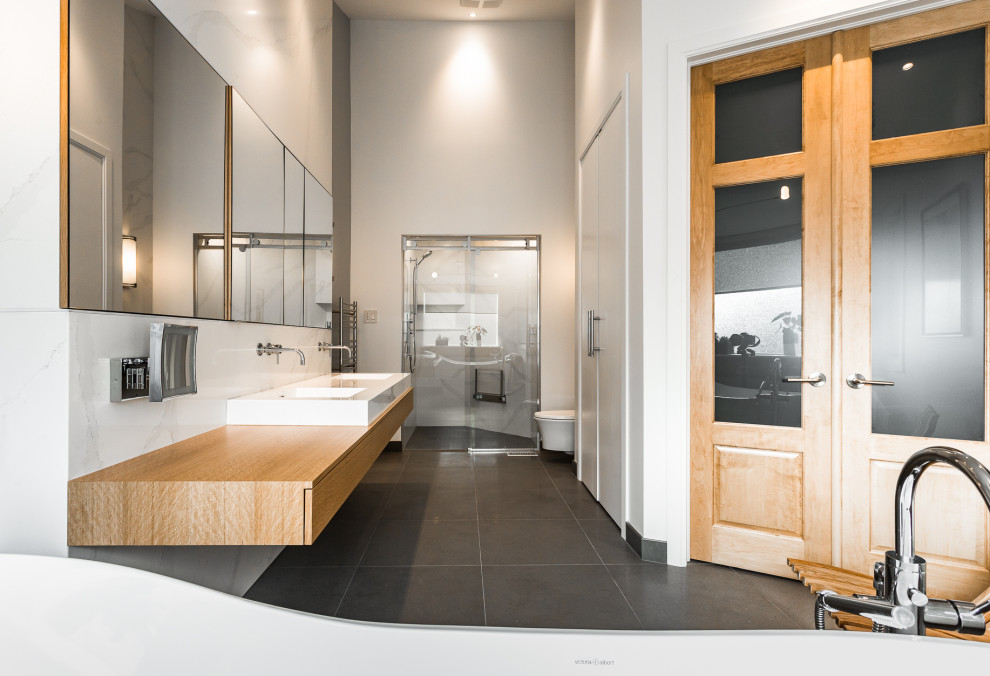 Photo of a large scandinavian master bathroom in Montreal with flat-panel cabinets, light wood cabinets, a freestanding tub, an alcove shower, a wall-mount toilet, white tile, stone slab, grey walls, ceramic floors, an integrated sink, solid surface benchtops, brown floor, a sliding shower screen, white benchtops, a niche, a double vanity, a floating vanity and vaulted.